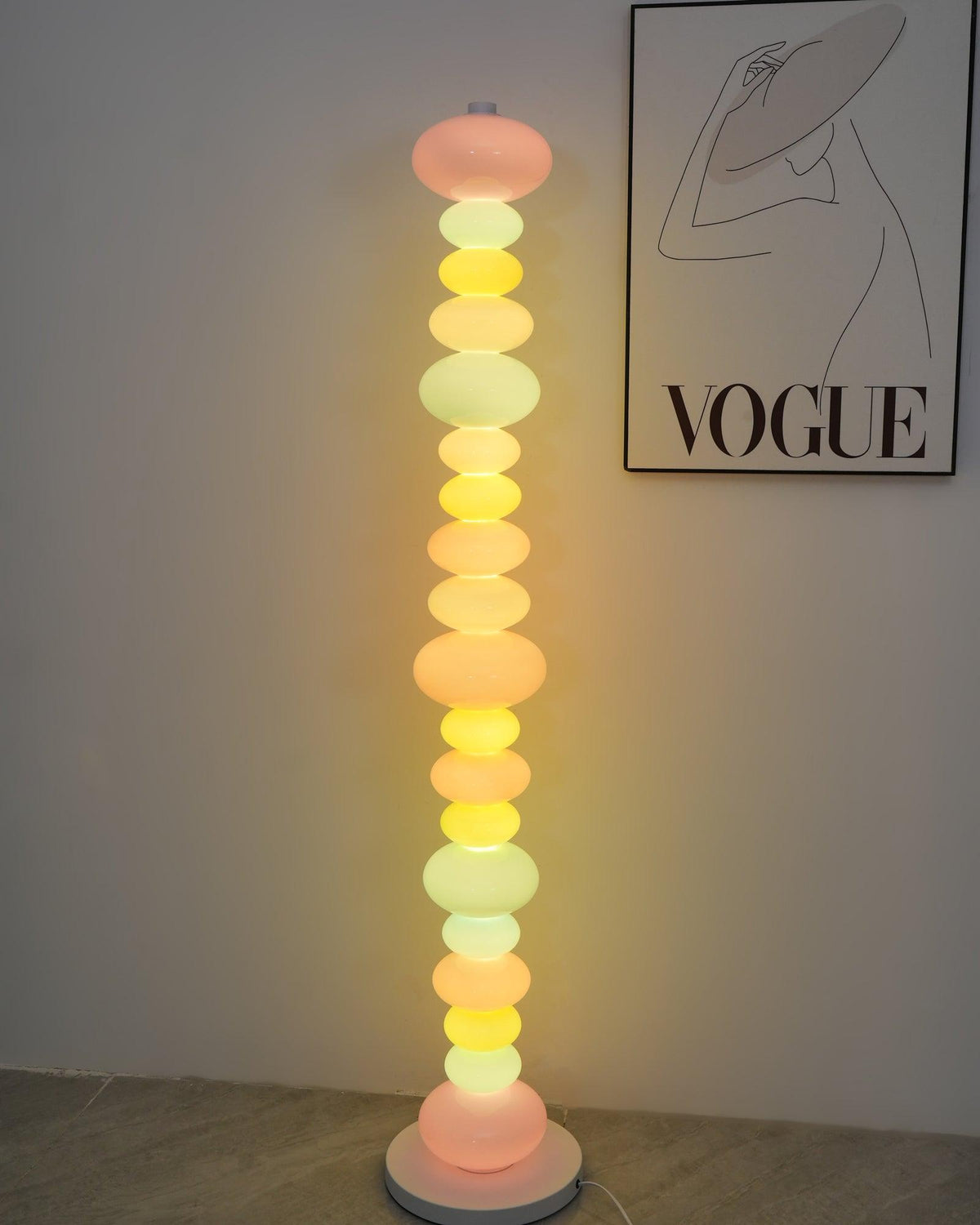 Clérachic stacked glass globe floor lamp lit in a narrow crop beside the same framed 'VOGUE' print on a white wall.