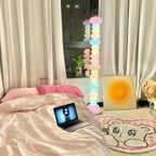 Clérachic multicolour floor lamp glowing in a bedroom with pink bedding, an open laptop and a bear-face rug.