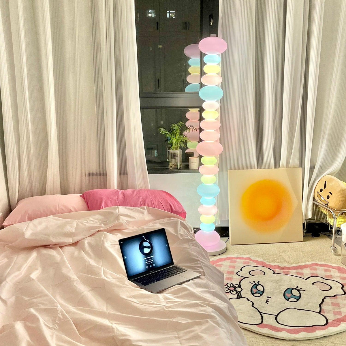 Clérachic multicolour floor lamp glowing in a bedroom with pink bedding, an open laptop and a bear-face rug.