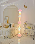 Clérachic multicolour floor lamp lit in a living room with a cream tufted sofa, a white cat resting on the floor nearby.