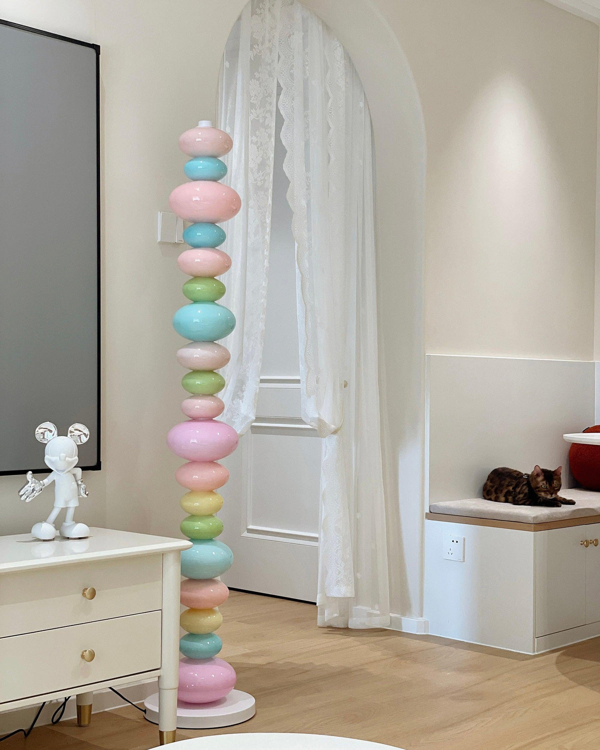A tall Clérachic floor lamp of stacked pastel glass globes stands lit beside a white nightstand in a softly lit bedroom corner.