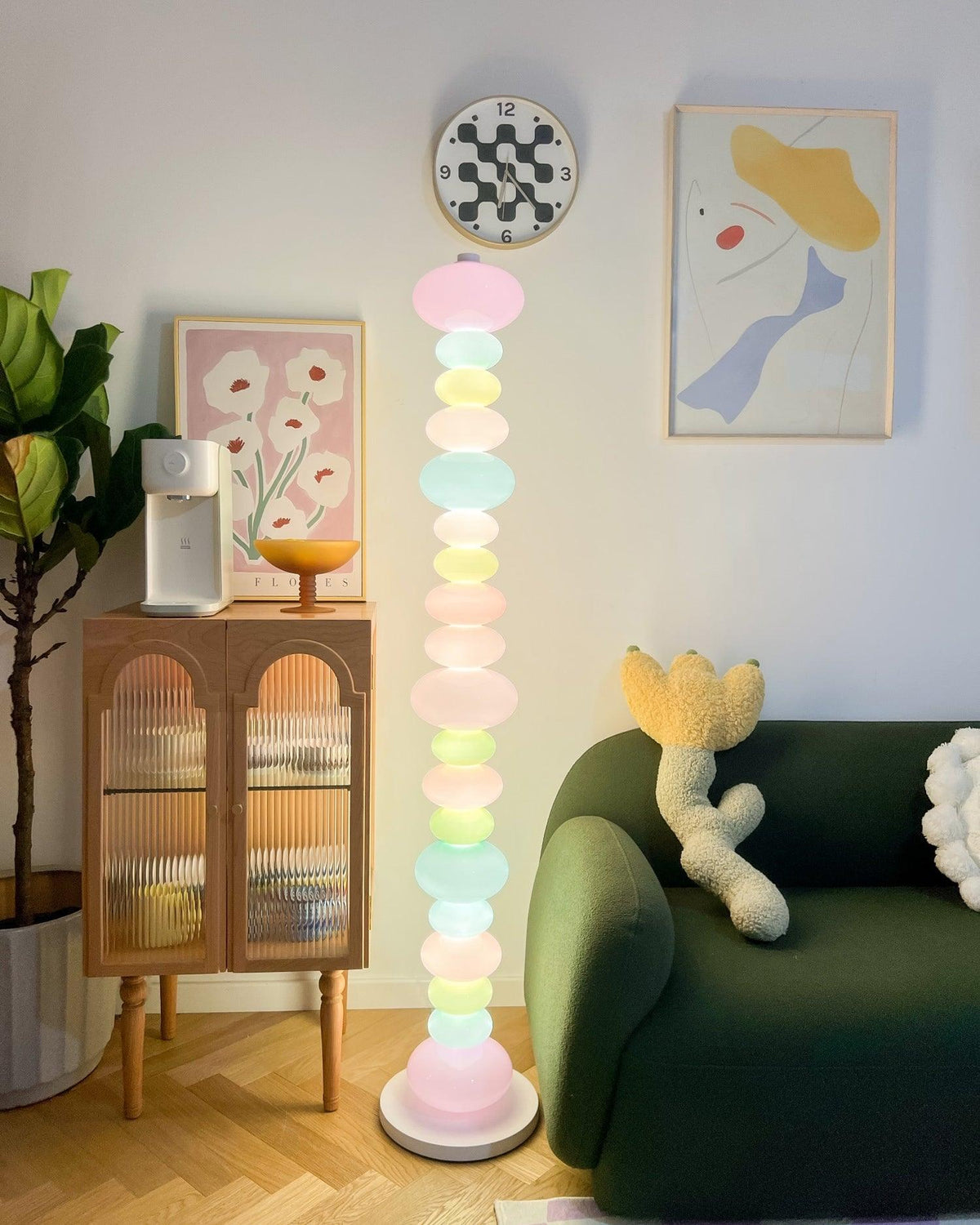Clérachic pastel glass globe floor lamp lit in a corner beside a green sofa, framed prints hung above on the wall.