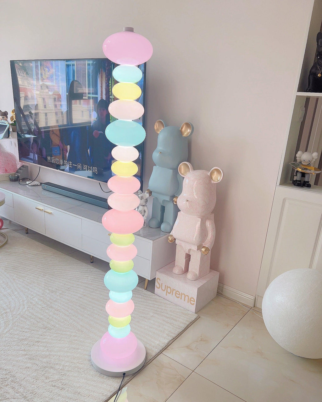 Clérachic multicolour floor lamp lit beside a television stand with oversized collectible bear figures on tiled floor.