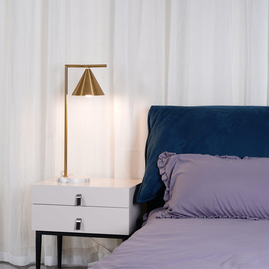 Lit and glowing warmly, the Clérachic lamp's cone shade is shown close-up beside a navy velvet headboard and lilac bedding.