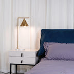 Lit and glowing warmly, the Clérachic lamp's cone shade is shown close-up beside a navy velvet headboard and lilac bedding.
