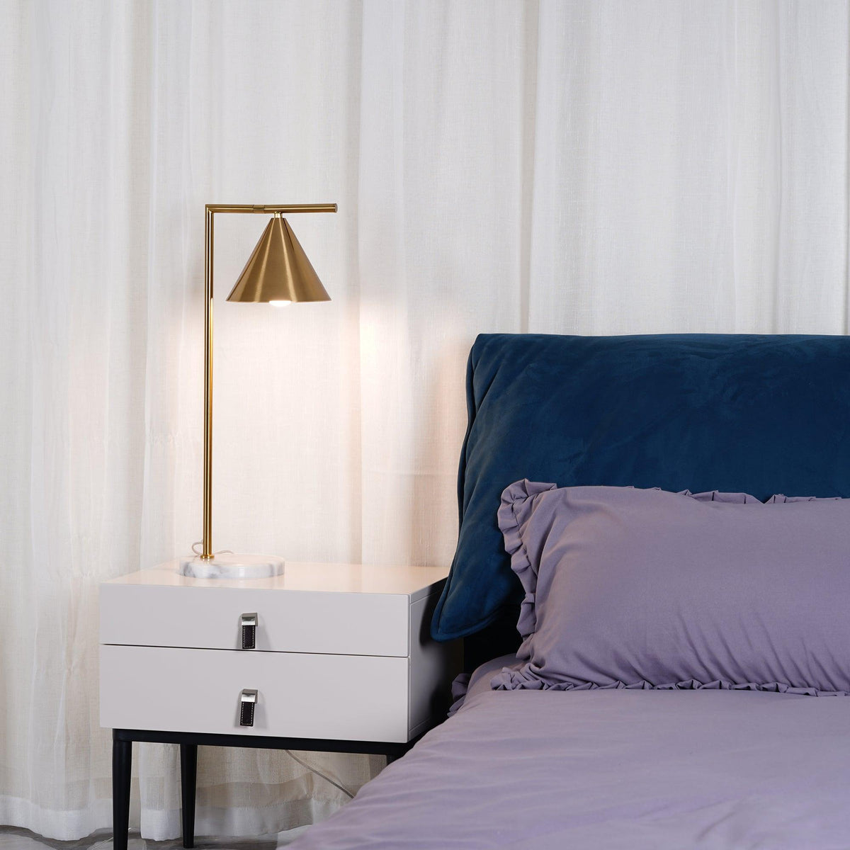 Lit and glowing warmly, the Clérachic lamp's cone shade is shown close-up beside a navy velvet headboard and lilac bedding.
