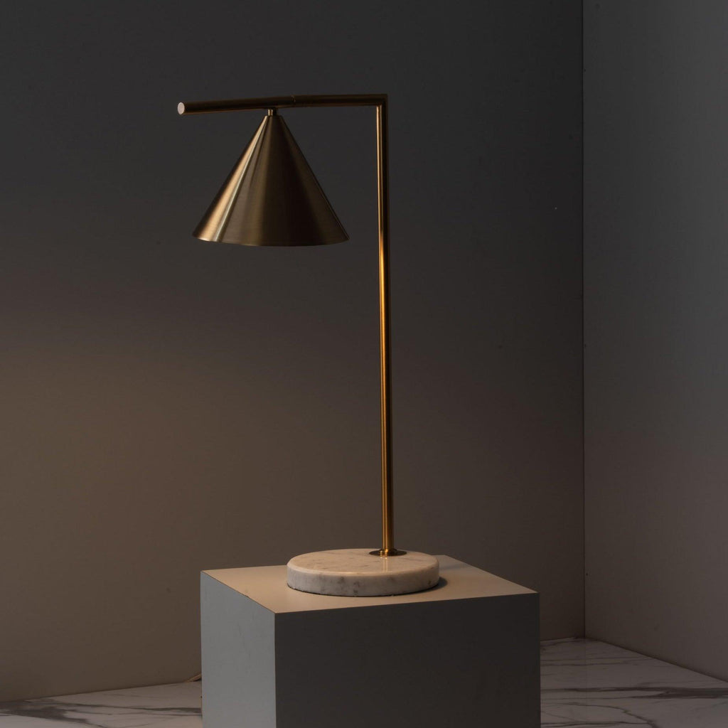Set on a grey stone plinth against a dark backdrop, this moody shot warms the Clérachic lamp's brass cone shade with amber light.