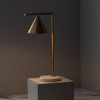 Set on a grey stone plinth against a dark backdrop, this moody shot warms the Clérachic lamp's brass cone shade with amber light.