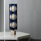 Three-globe Clérachic lamp in dark navy on a warm stone base, standing on a white marble side table by a window.
