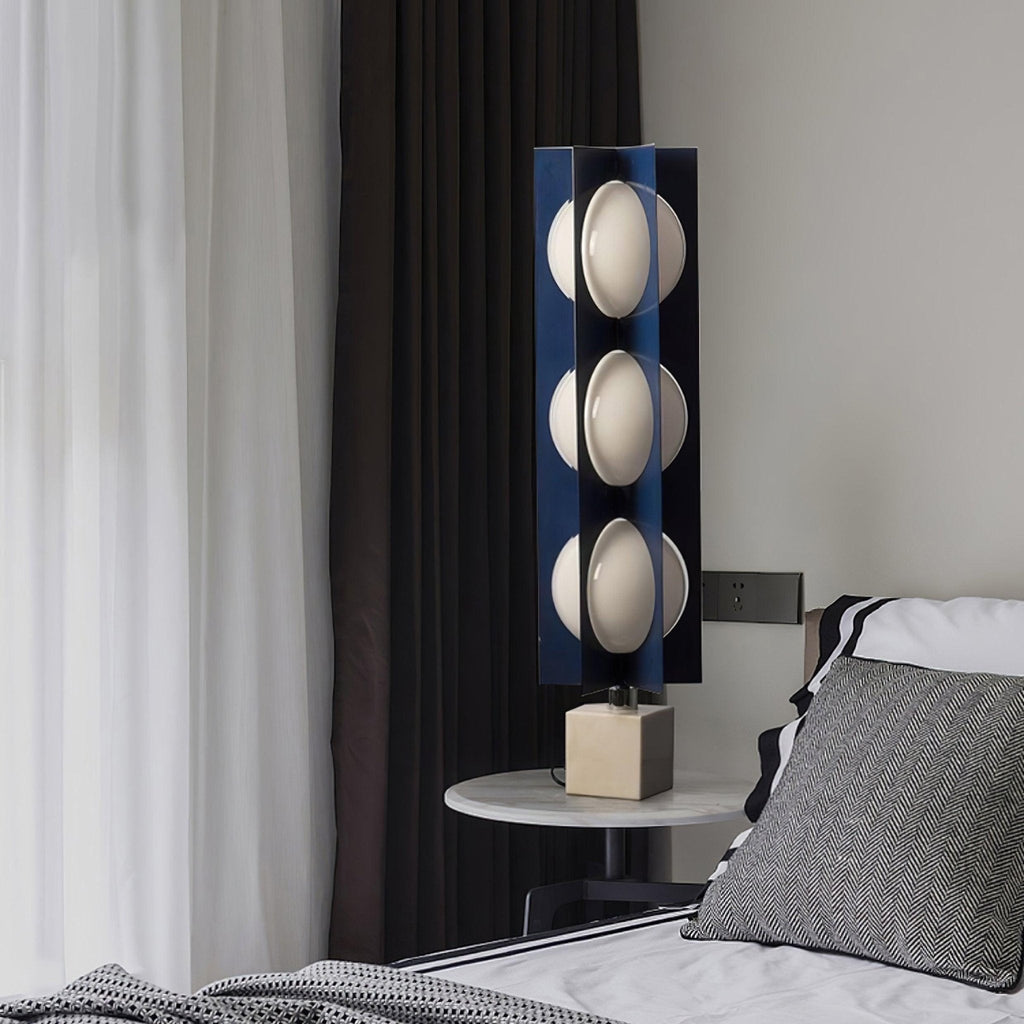 Three-globe Clérachic lamp glowing on a round bedside table, beside a striped pillow and a dark bedroom curtain.