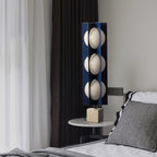 Three-globe Clérachic lamp glowing on a round bedside table, beside a striped pillow and a dark bedroom curtain.