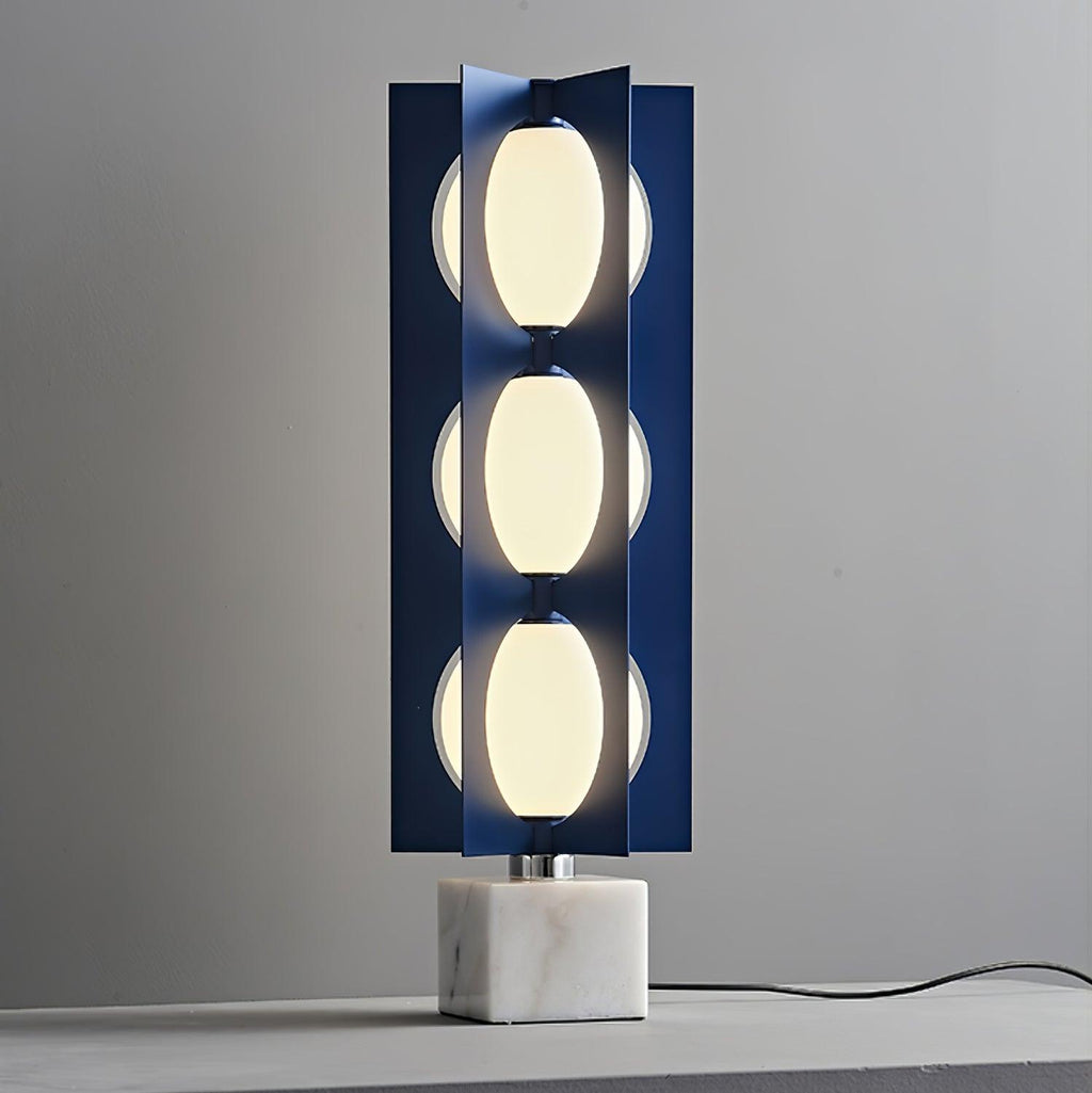 Three-globe Clérachic lamp lit on a tabletop, its cord trailing across a plain grey surface in a bare studio set.