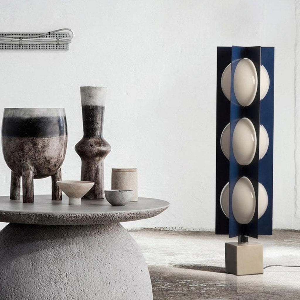Three-globe Clérachic floor lamp glowing beside a cluster of dark ceramic vessels on a round stone side table.