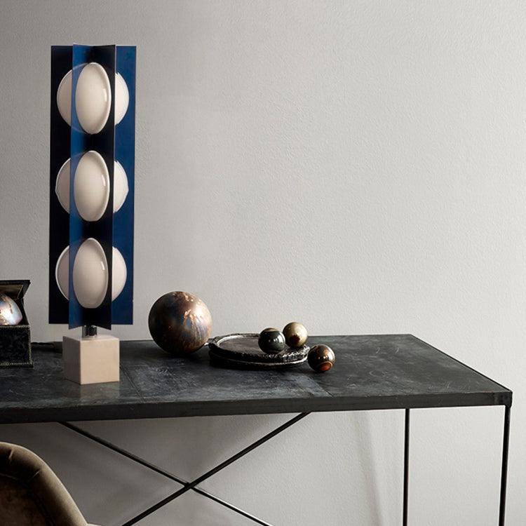 Three-globe Clérachic lamp on a warm stone base, set on a dark marble console beside small metallic spheres.