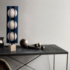 Three-globe Clérachic lamp on a warm stone base, set on a dark marble console beside small metallic spheres.
