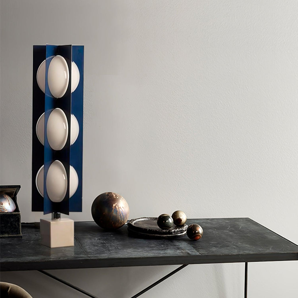 Three-globe Clérachic lamp on a warm stone base, standing on a dark console table beside a bowl of metallic spheres.