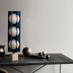 Three-globe Clérachic lamp on a warm stone base, standing on a dark console table beside a bowl of metallic spheres.