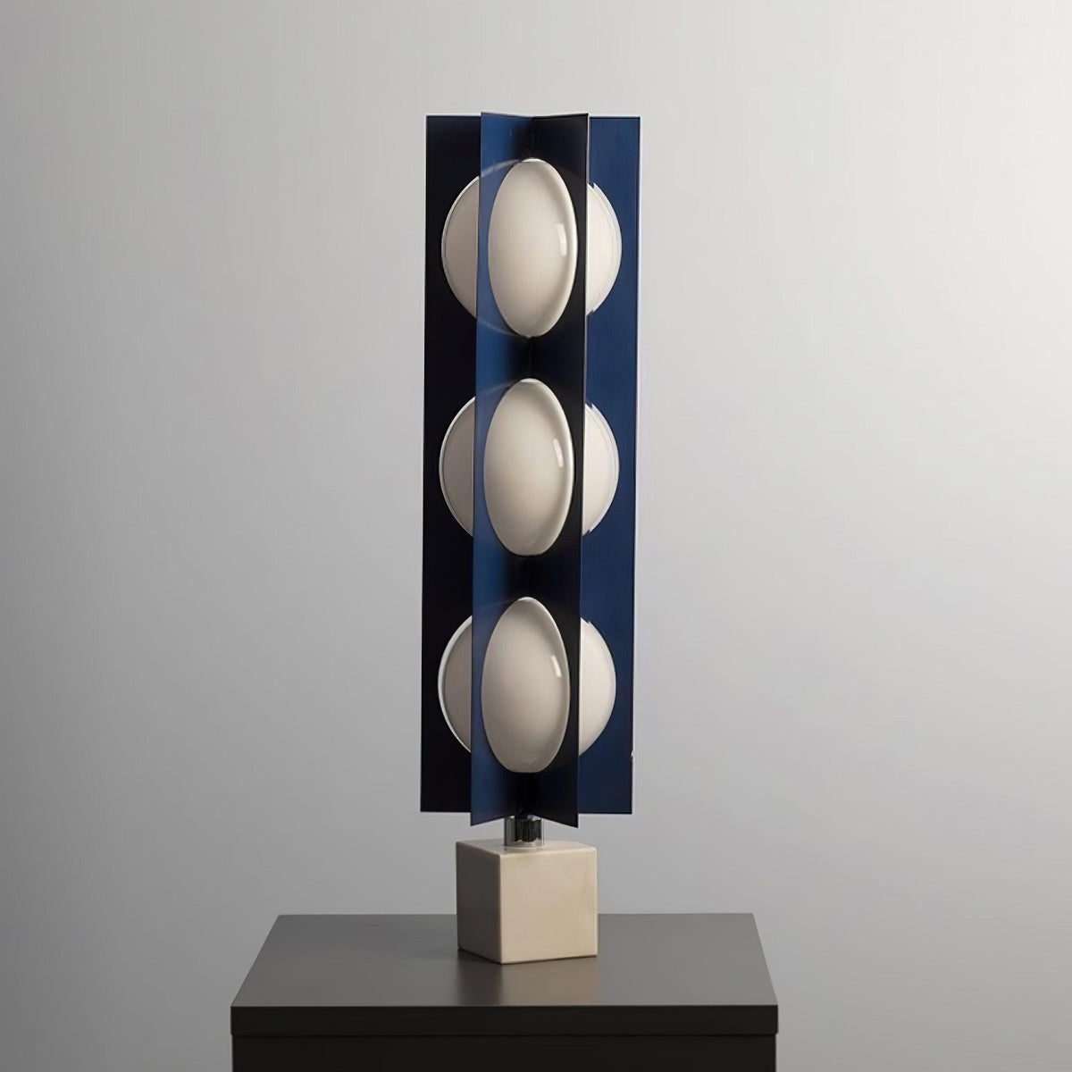 Three-globe Clérachic lamp on a warm stone base, standing on a dark wood nightstand against a plain grey wall.