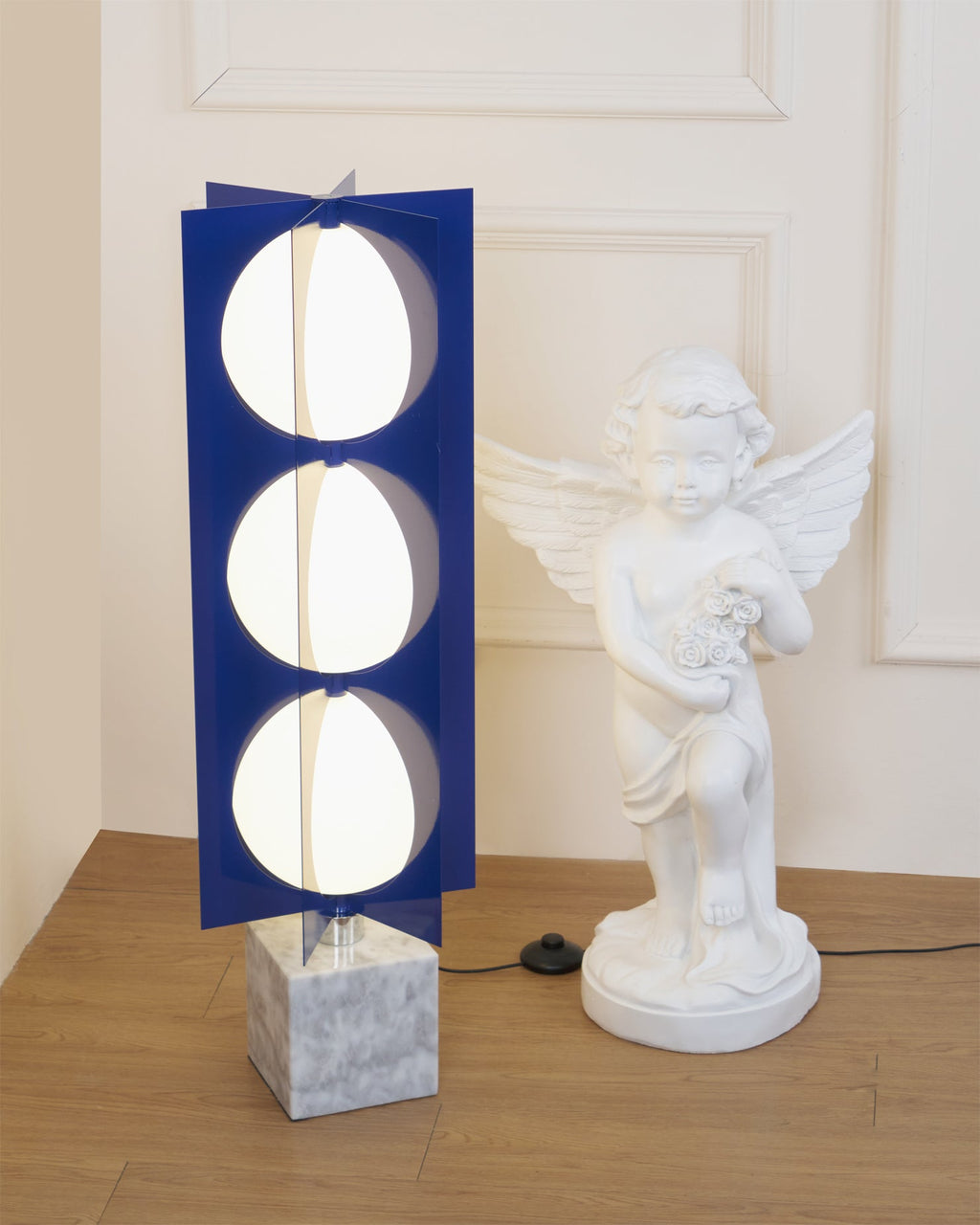 Three-globe Clérachic lamp lit beside a cherub statue, framed within a panelled doorway in a warm-toned room.