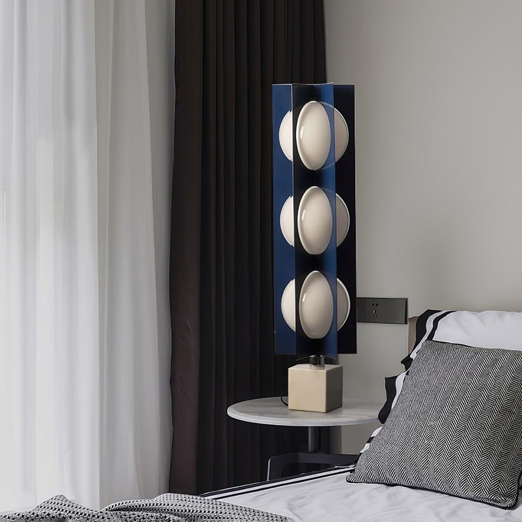 Three-globe Clérachic table-height lamp glowing beside a bed, its light falling across a striped pillow and dark curtain.