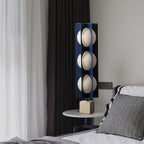 Three-globe Clérachic table-height lamp glowing beside a bed, its light falling across a striped pillow and dark curtain.