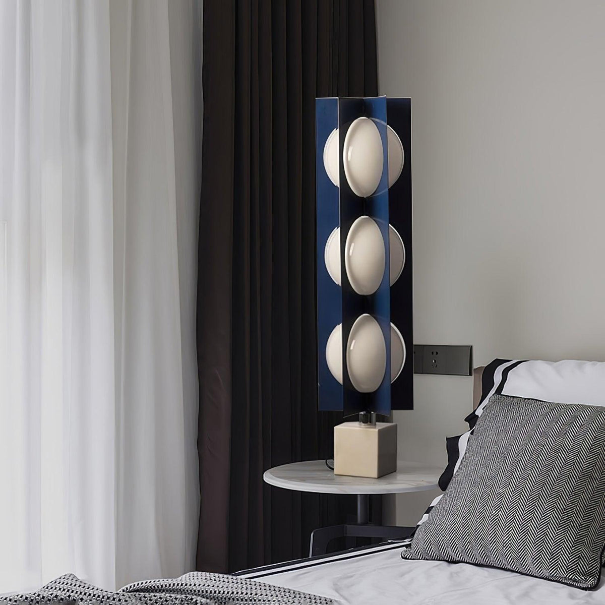 Three-globe Clérachic table-height lamp glowing beside a bed, its light falling across a striped pillow and dark curtain.