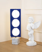 Three-globe Clérachic floor lamp on a white marble base, framed close beside a cherub statue on a timber floor.