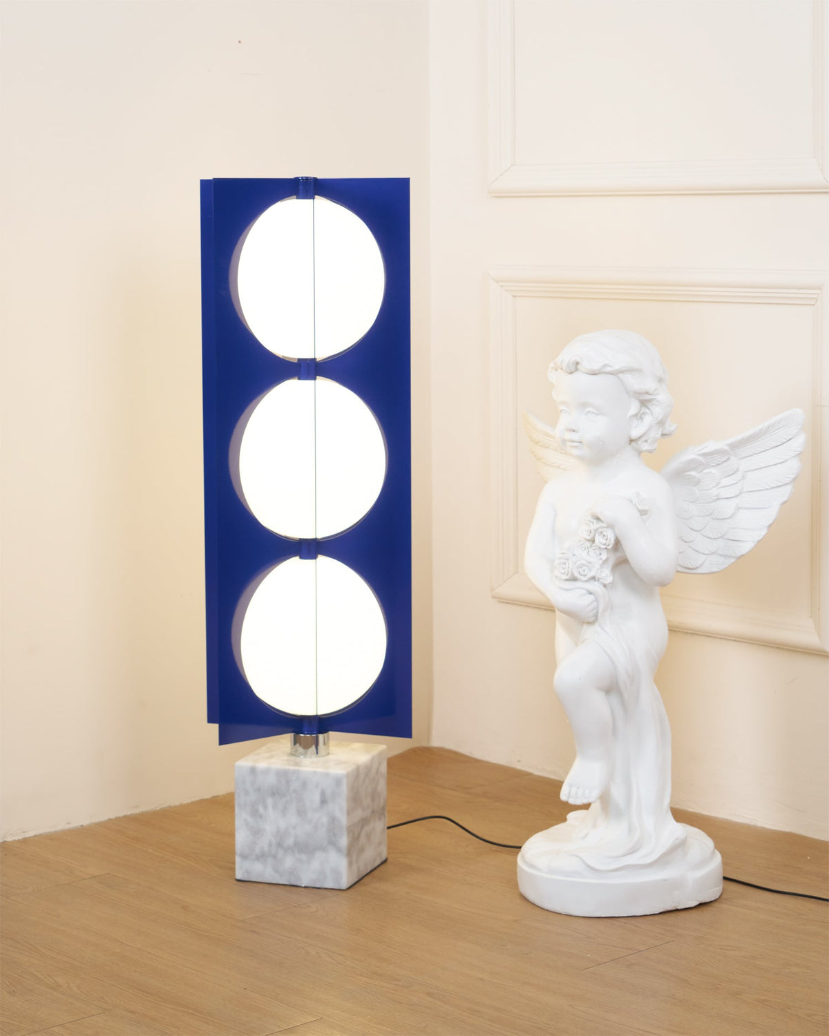 Three-globe Clérachic floor lamp on a white marble base, framed close beside a cherub statue on a timber floor.