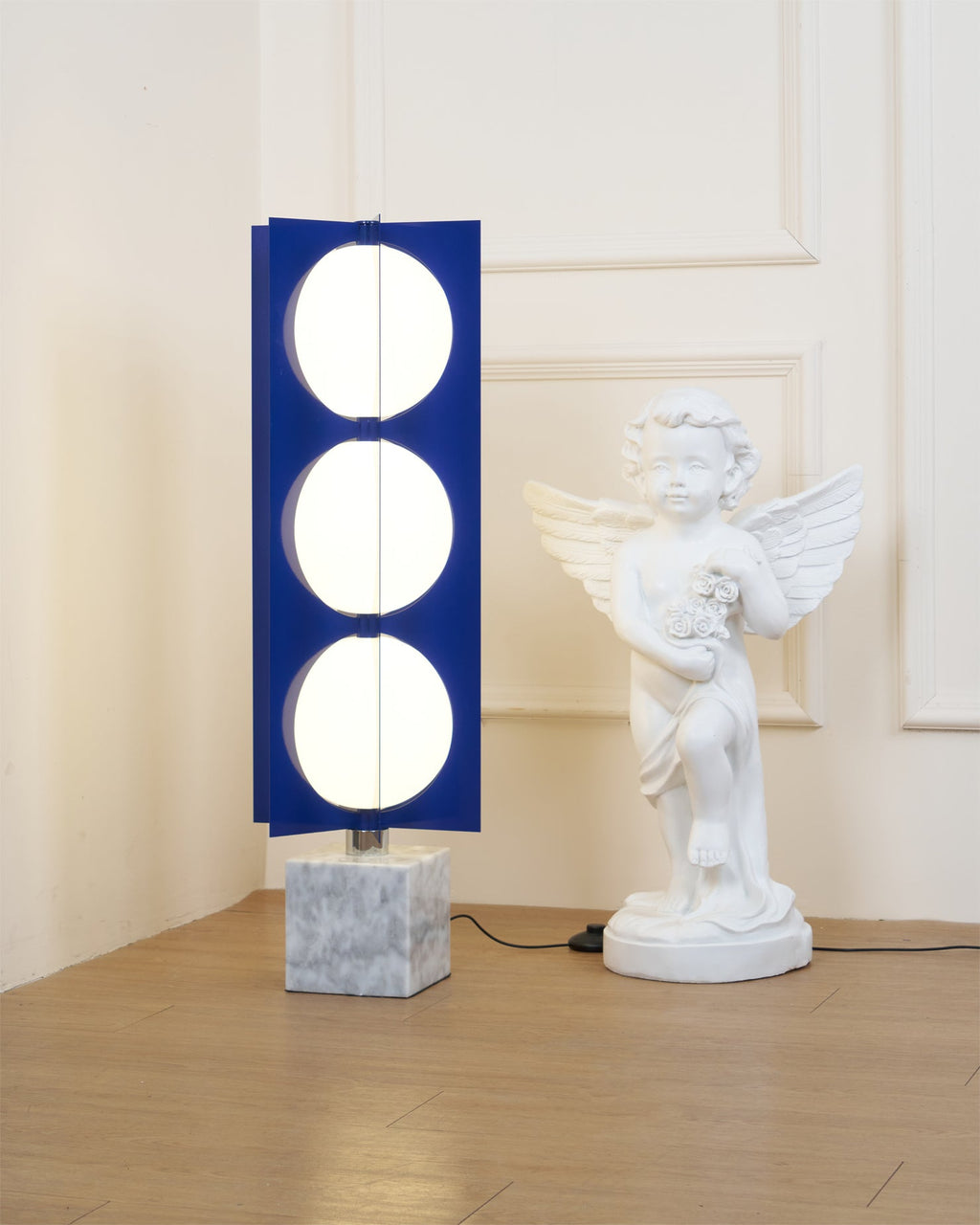 Three-globe Clérachic floor lamp on a white marble base, standing beside a cherub statue in a sunlit room corner.