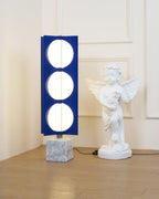 Three-globe Clérachic floor lamp on a white marble base, standing beside a cherub statue in a sunlit room corner.