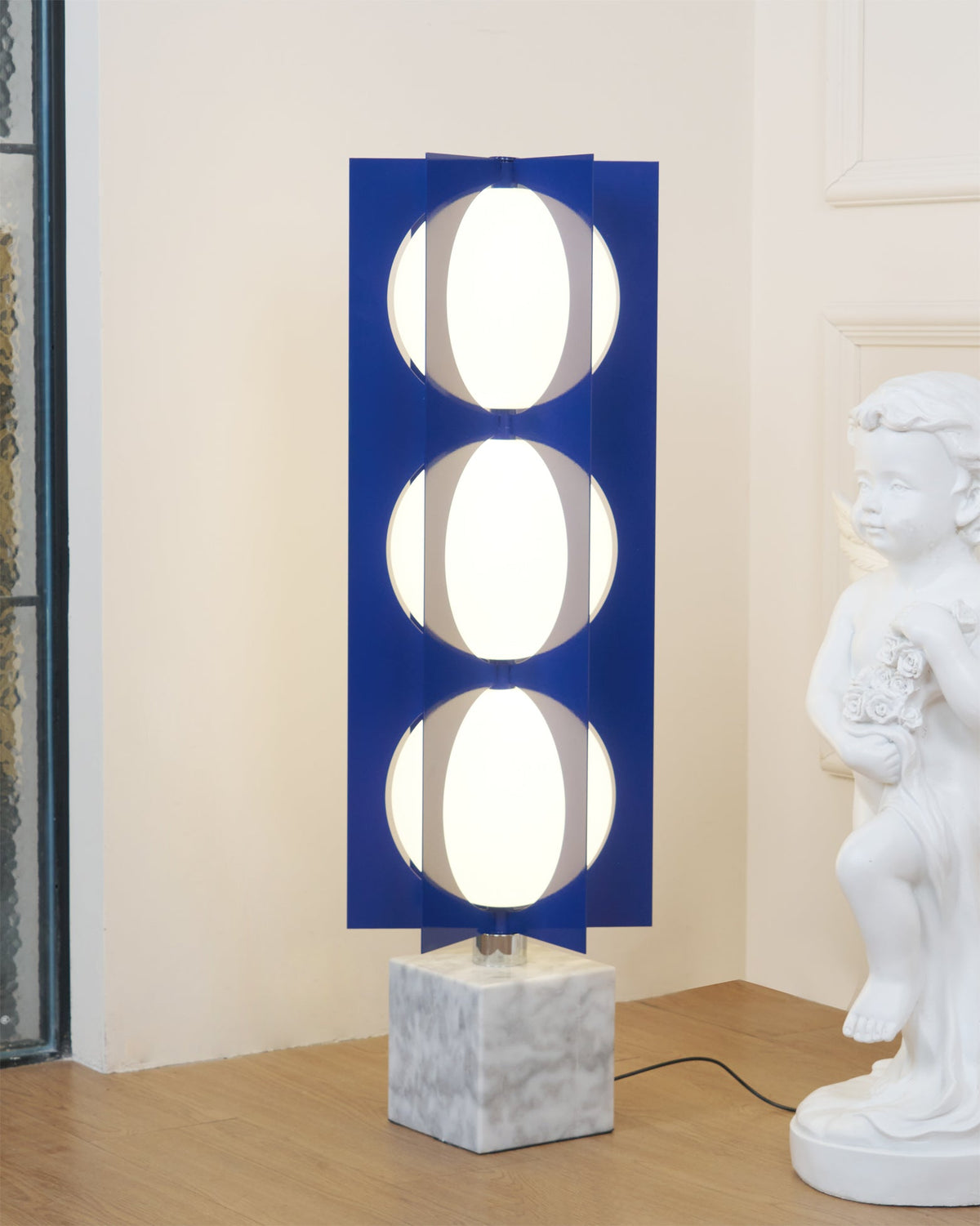 Three-globe Clérachic floor lamp glowing beside a cherub statue, set against a panelled wall and timber floor.