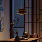 In a dim evening room, this Clérachic pendant in walnut wood hangs beside a paper screen and a dark wood table holding a branch in a vase.