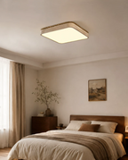 This square Clérachic ceiling light glows warm above a bed dressed in soft neutral linens, beside a sheer-curtained window.