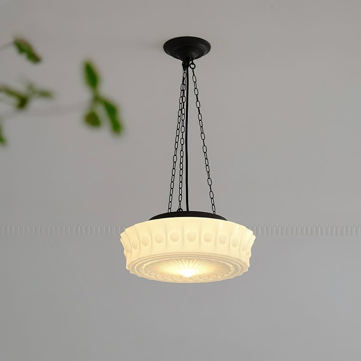 A close hanging view of this beige fluted-glass Clérachic pendant, lit, with a blurred green leaf at the edge of the frame.