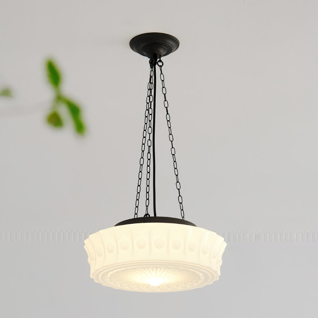 A close angled view of this beige fluted-glass Clérachic pendant, lit, with blurred green leaves in the foreground.
