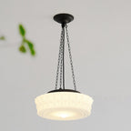 A close angled view of this beige fluted-glass Clérachic pendant, lit, with blurred green leaves in the foreground.