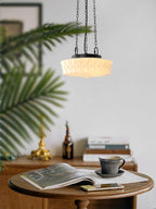 Hanging low over a round wooden table set with a magazine and coffee cup, this beige fluted-glass Clérachic pendant glows.