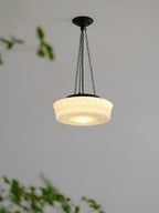 A close hanging view of this beige fluted-glass Clérachic pendant, lit, with blurred green leaves crossing the frame.