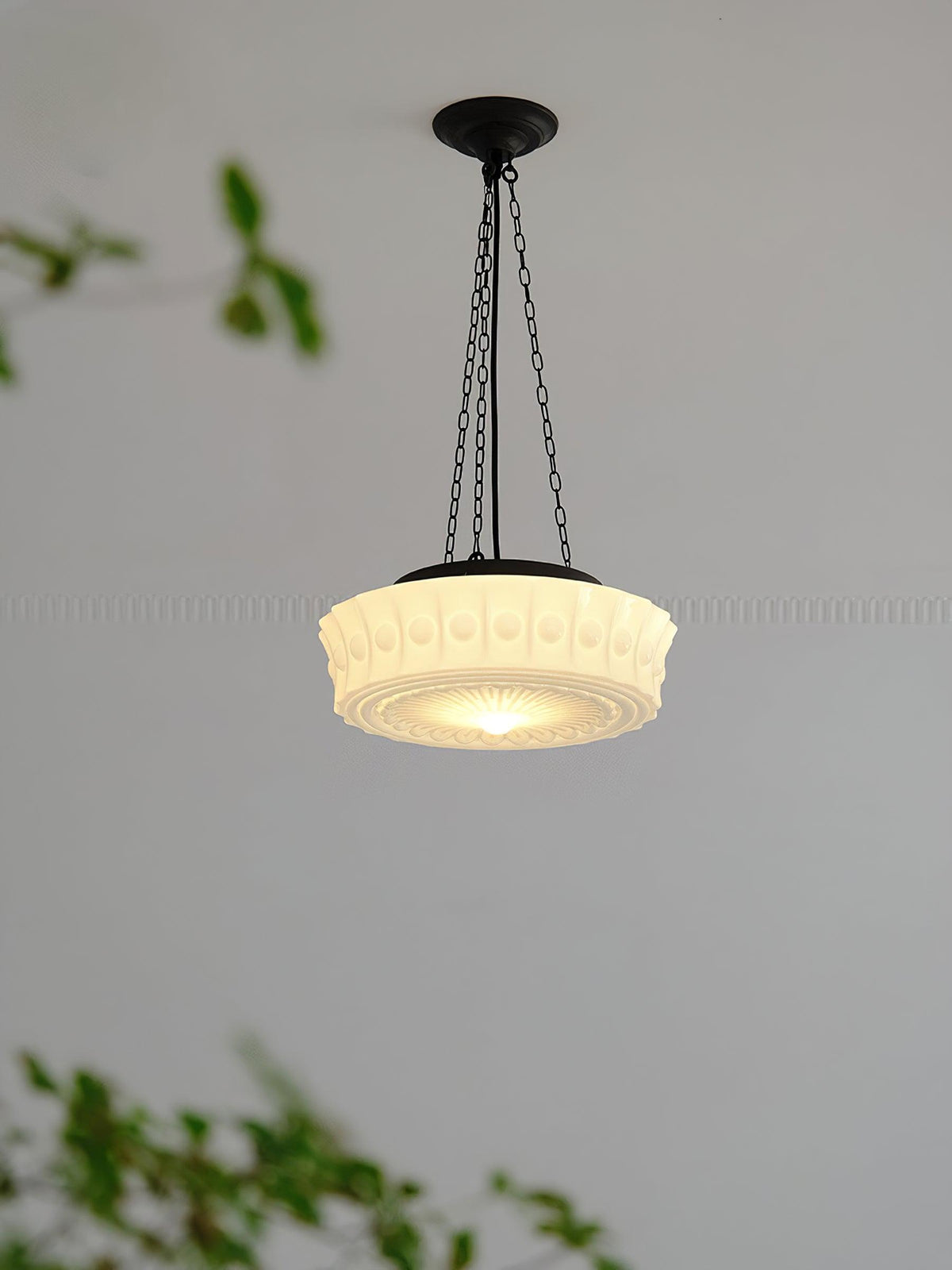 A close hanging view of this beige fluted-glass Clérachic pendant, lit, with blurred green leaves crossing the frame.