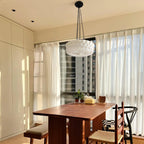 Beside sheer curtains with a city view outside, this beige fluted-glass Clérachic pendant hangs above a wood dining table.