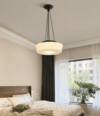 Over a bed with pillows, this beige fluted-glass Clérachic pendant hangs with a green curtain framing the window behind it.