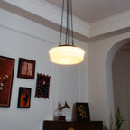 Near an archway with framed pictures and a gramophone horn on a dark wood cabinet, this beige fluted-glass Clérachic pendant hangs.