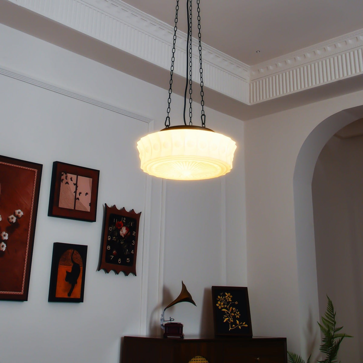 Near an archway with framed pictures and a gramophone horn on a dark wood cabinet, this beige fluted-glass Clérachic pendant hangs.