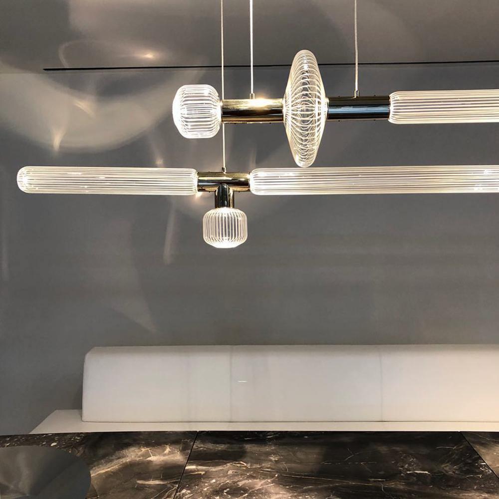 A linear Clérachic pendant with fluted glass cylinders and a round disc hangs lit above a white bench.