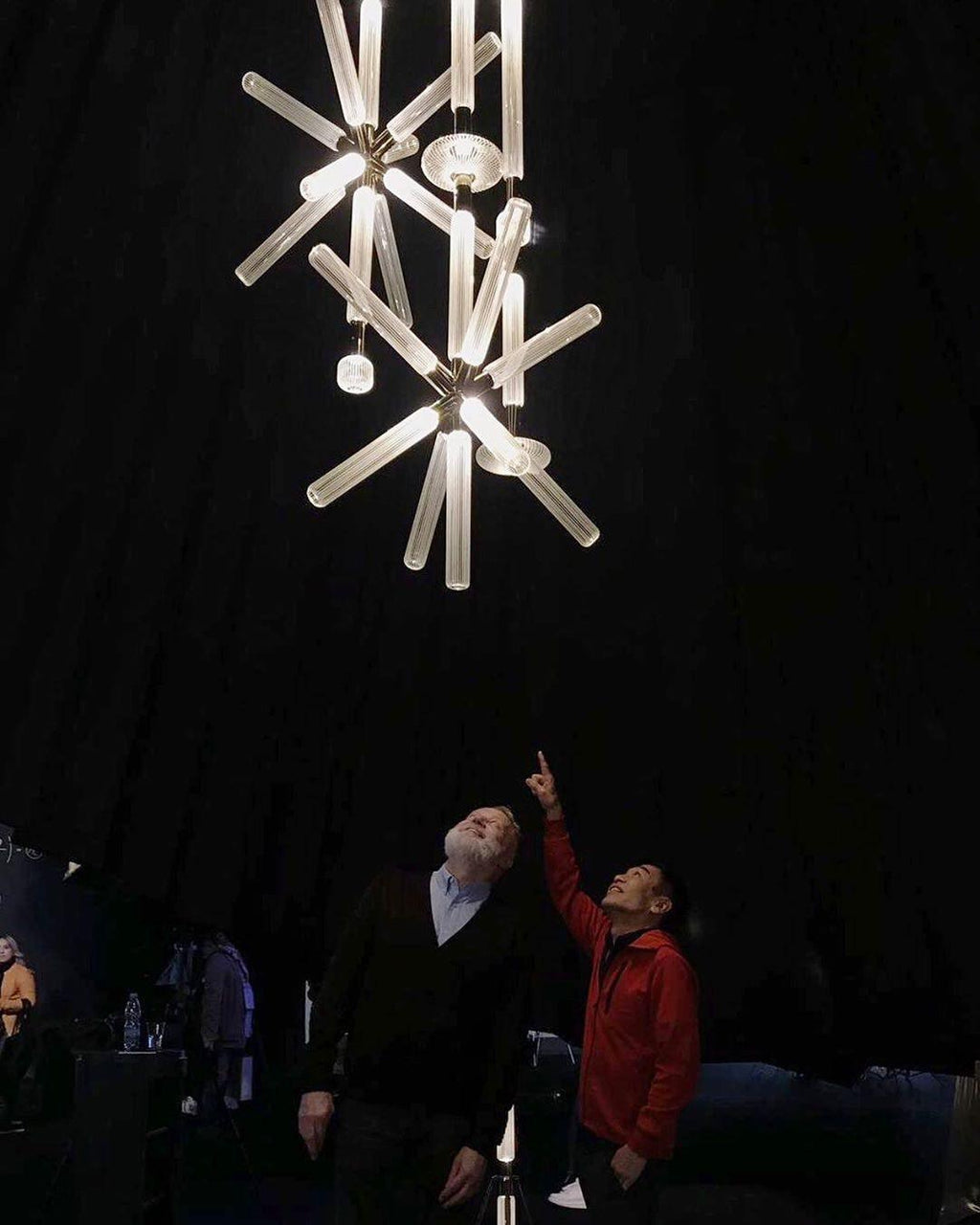 People look up at a lit Clérachic pendant cluster hanging high overhead in a dark venue.