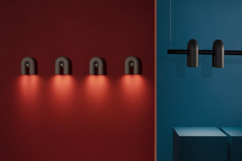 Four black Clérachic wall lights glow in a row on a red wall, beside spotlights mounted on a rod against teal.