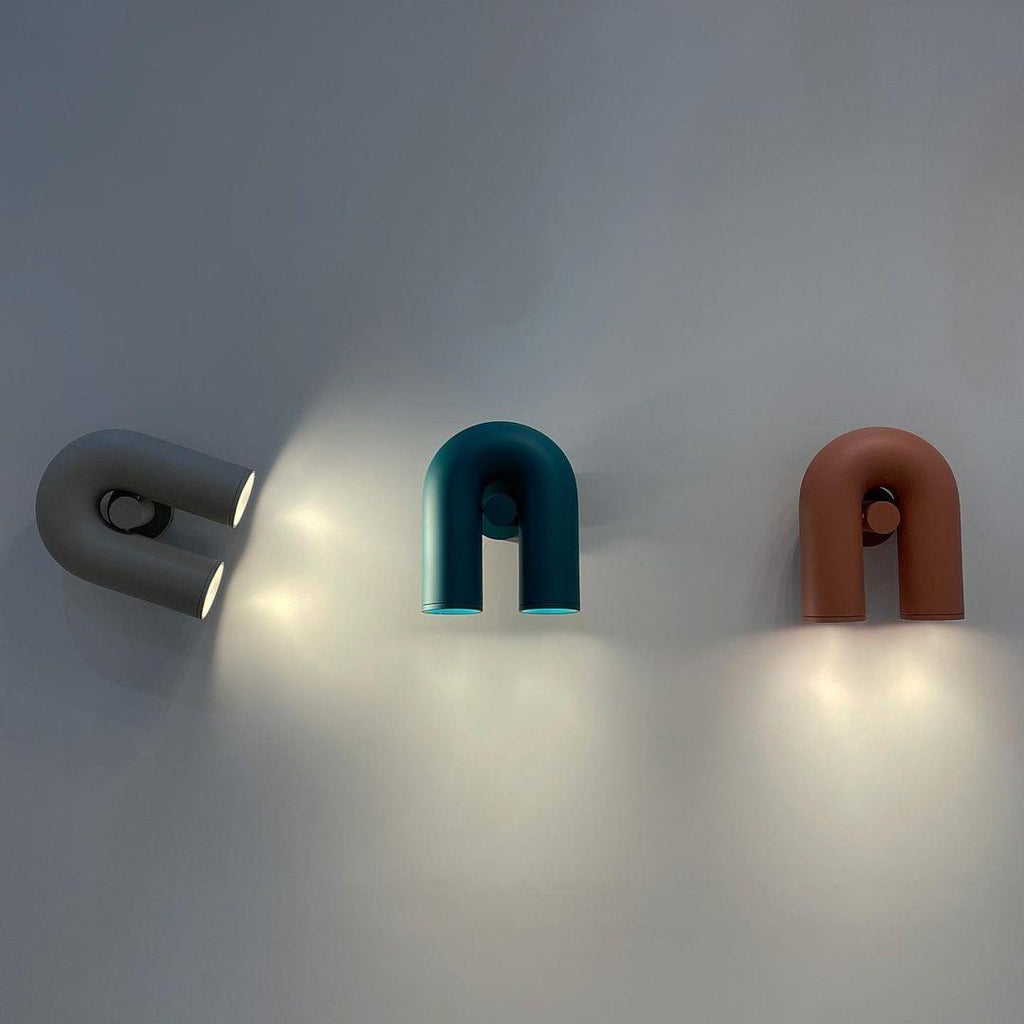 Two Clérachic wall lights, one teal and one terracotta, glow beside a swivelling spotlight on a plain grey wall.