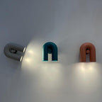 Two Clérachic wall lights, one teal and one terracotta, glow beside a swivelling spotlight on a plain grey wall.