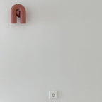 This Clérachic wall light in a dusty-pink finish mounts high on a plain wall above an electrical socket, unlit.