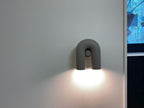 This charcoal Clérachic wall light casts bright white light on a wall beside a daylit window.